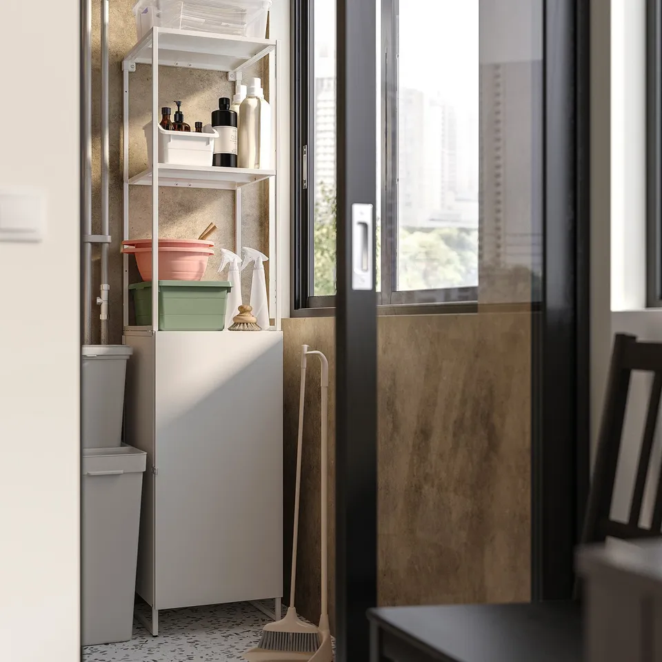 A JOSTEIN storage unit in a room. Its white, made of steel, and has multiple shelves holding cleaning supplies.