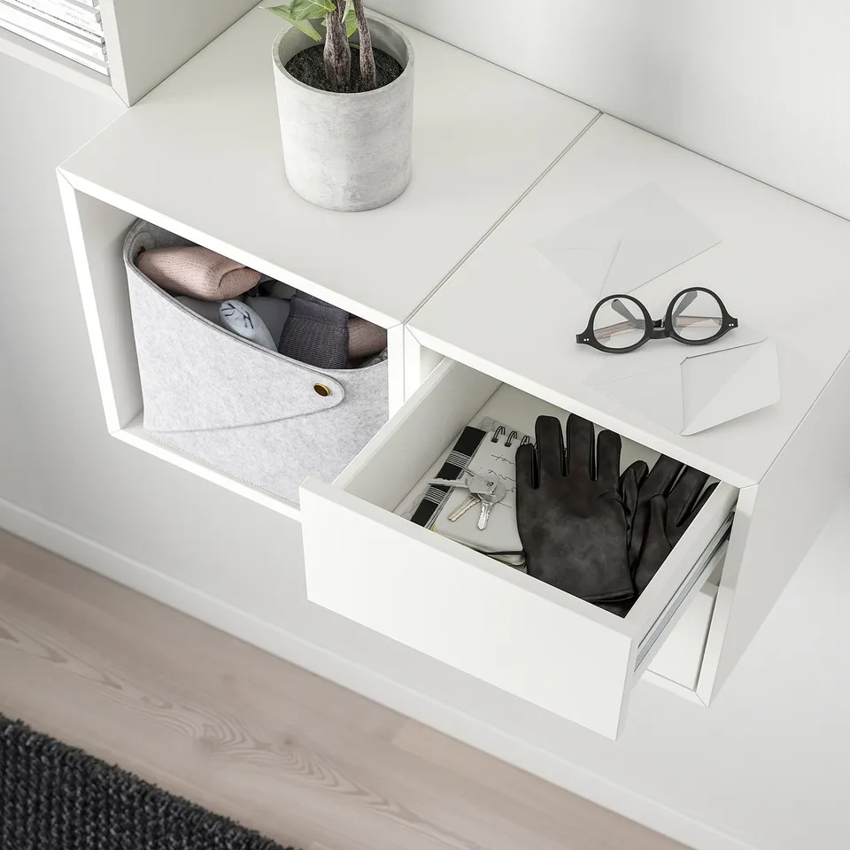 White wall-mounted storage unit with open compartments and one open drawer showing gloves, keys, and a notepad. A potted plant sits on top.