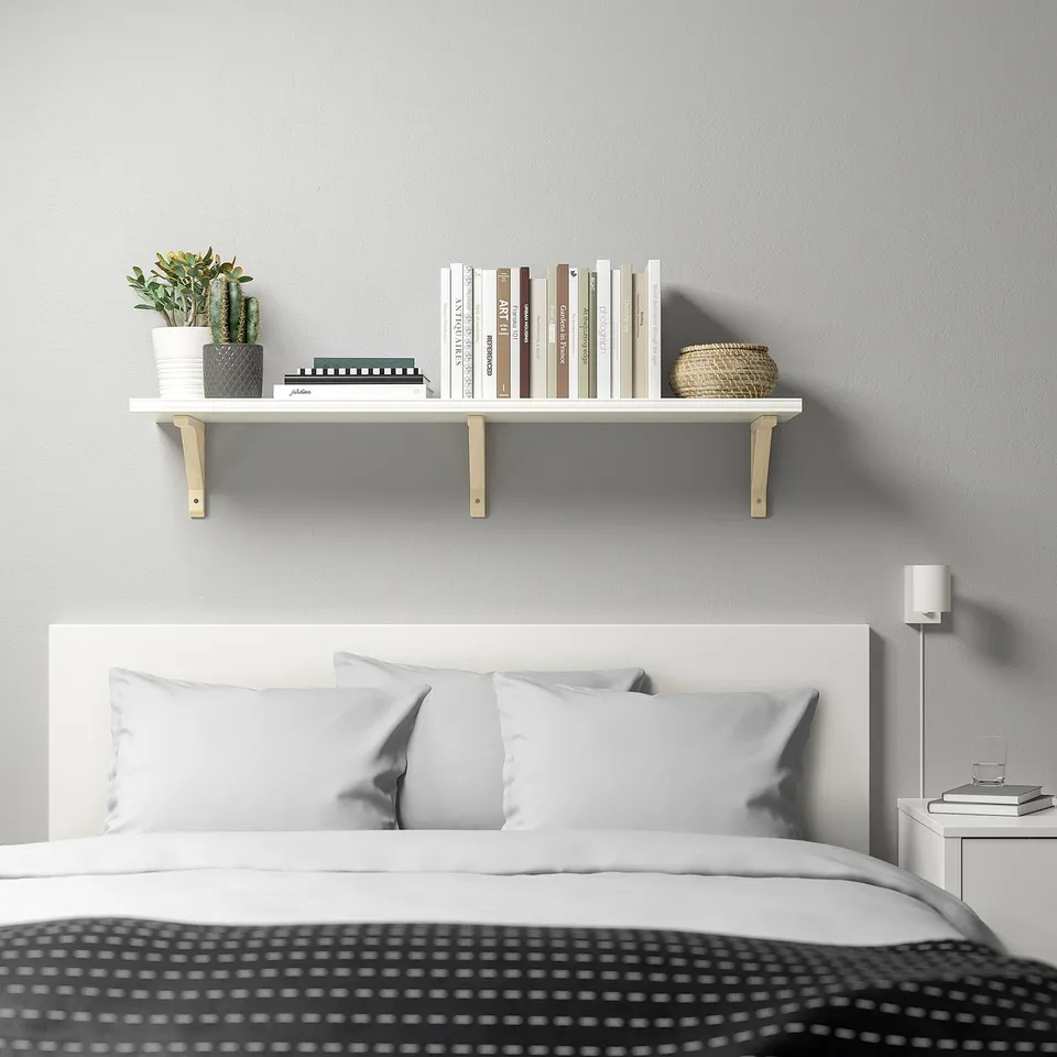 BERGSHULT white wall shelf, wooden brackets, holds books, plants. Mounted above bed, near reading lamp. Shelf is narrow, long.