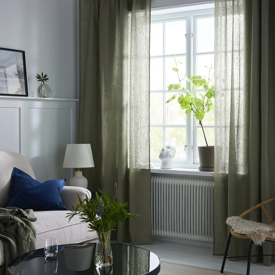 Modern living room with large olive green linen curtains behind a cosy chair with blue and white pillows, a white lamp, and a vase with greenery.