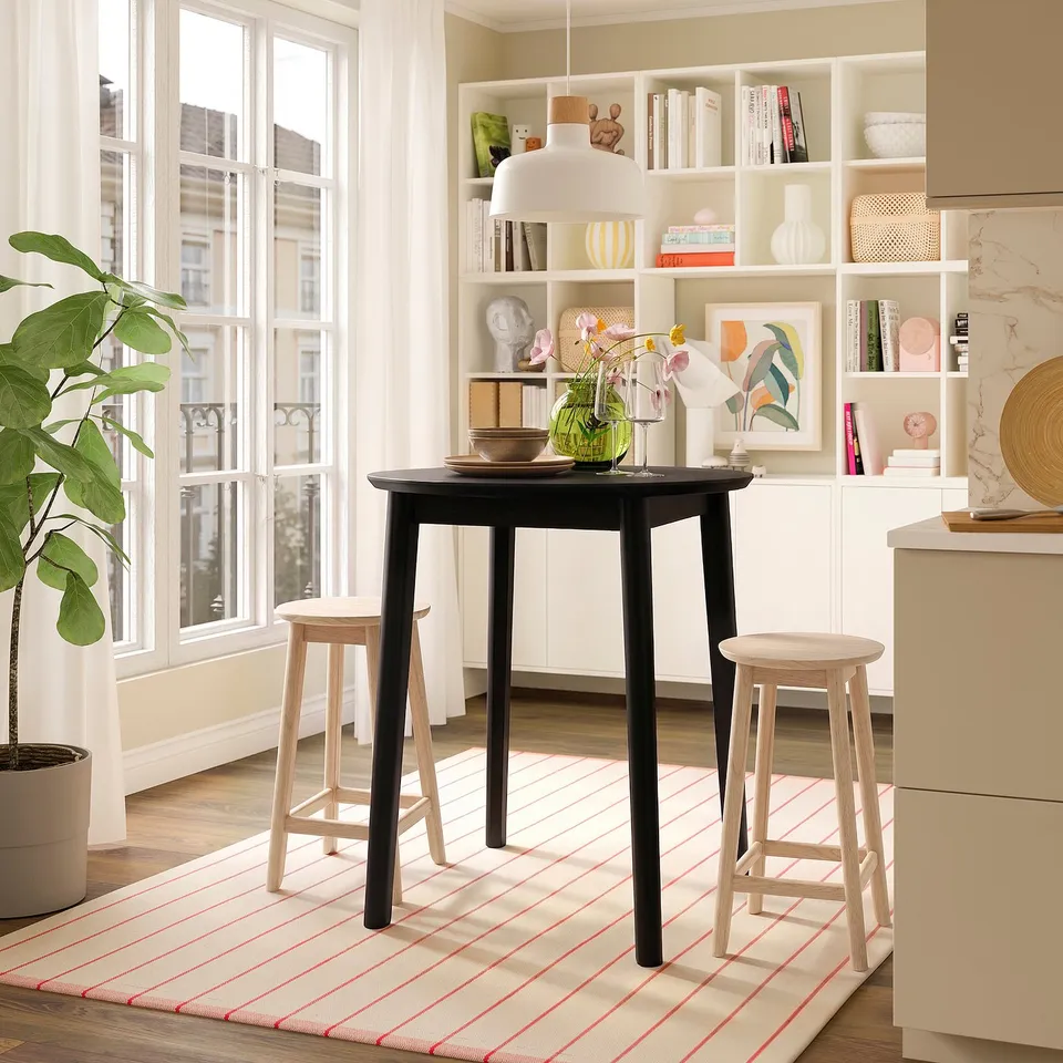 Modern dining: RÄVSTEN dark wood table with light stools, rug, window, and bookshelf.
