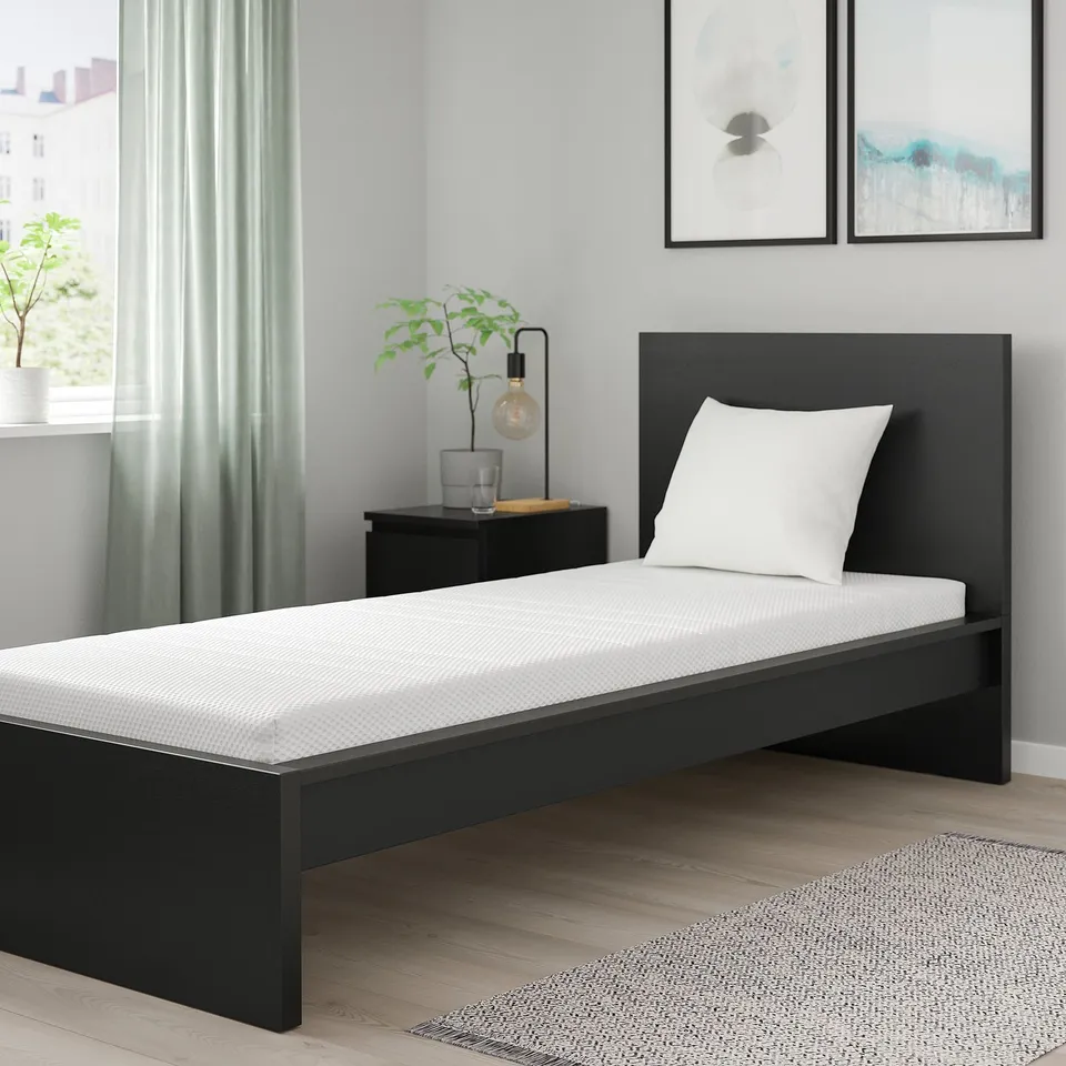 Modern bedroom with black ÅKREHAMN mattress, single pillow, abstract art, green curtain, bedside lamp, plant, patterned rug.