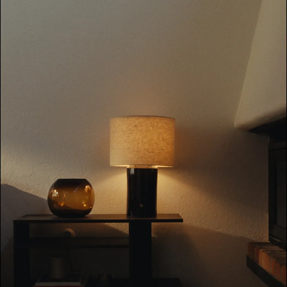 Table lamp HALGATT, with beige linen shade and ceramic base, sits on dark surface. Soft warm light glows, creating cosy ambiance.