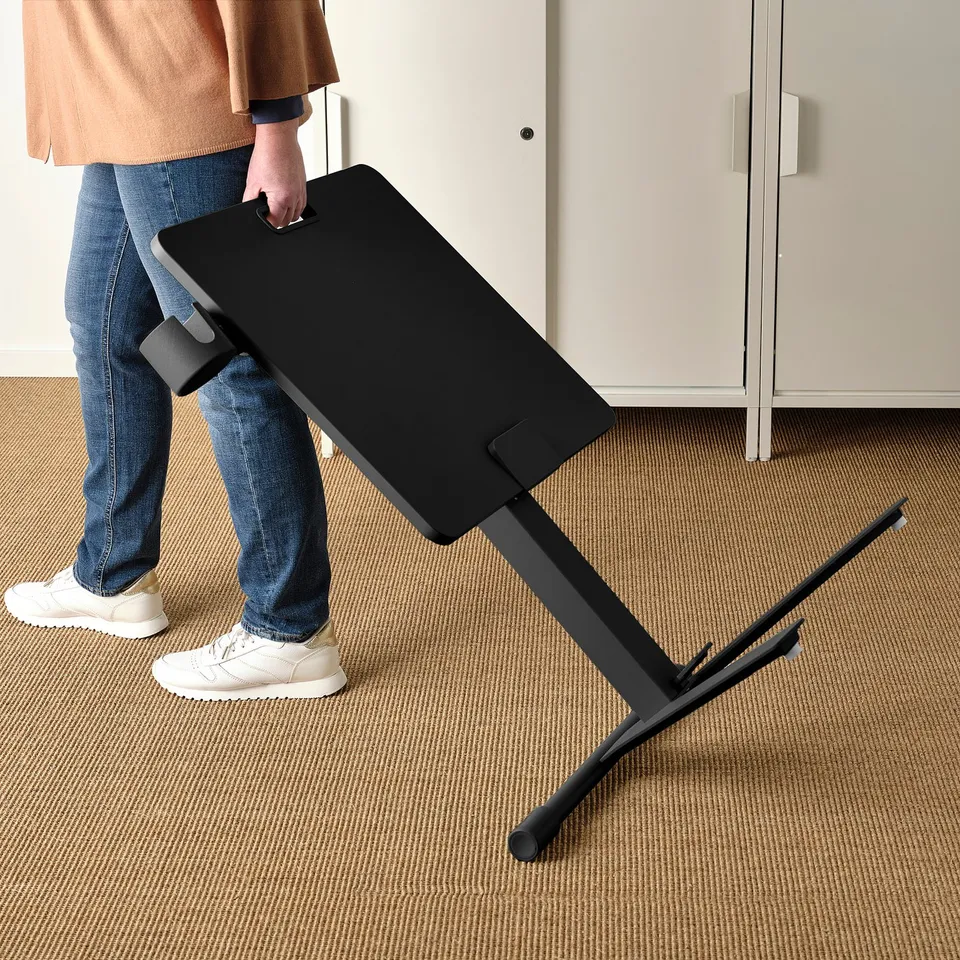 A person adjusting the height of a black MITTZON laptop table on a carpeted floor. The table has a tiltable top and castors for mobility.