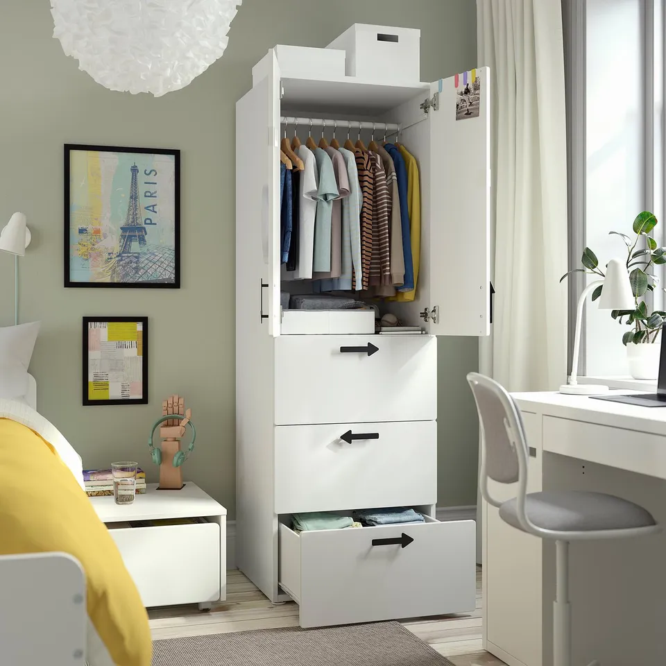 White wardrobe with clothes, drawers, and a box on top in a bedroom. Decorated with framed paris poster, lamp, and plant.