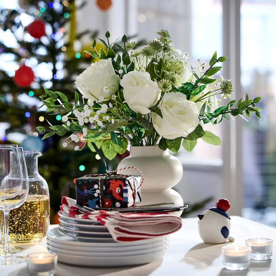 Christmas table with white roses, plates, glasses, VINTERFINT gift wrap, and snowman candle. Background has decorated tree.