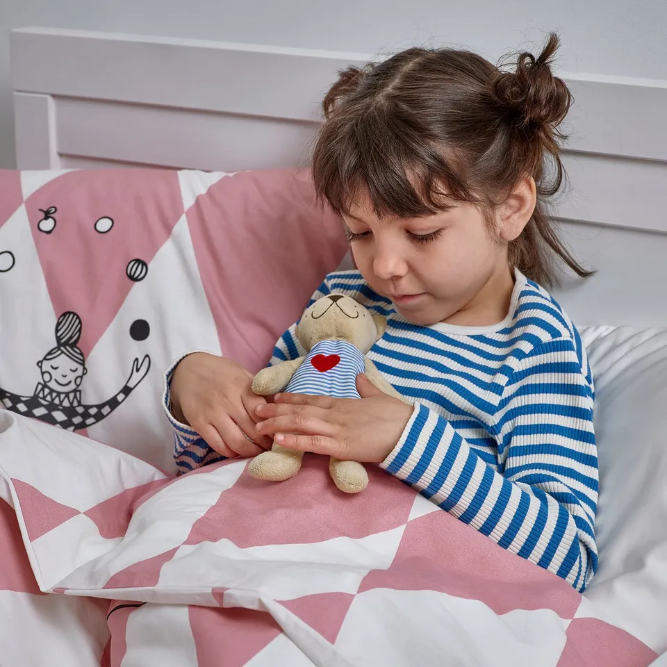 Girl in striped pyjamas sits on bed, holding heart teddy bear.