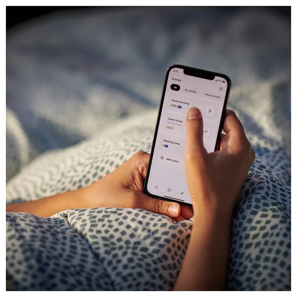 A persons hand holds a smartphone displaying the IKEA home smart app with lighting scenes. The person wears patterned blue pyjama pants.