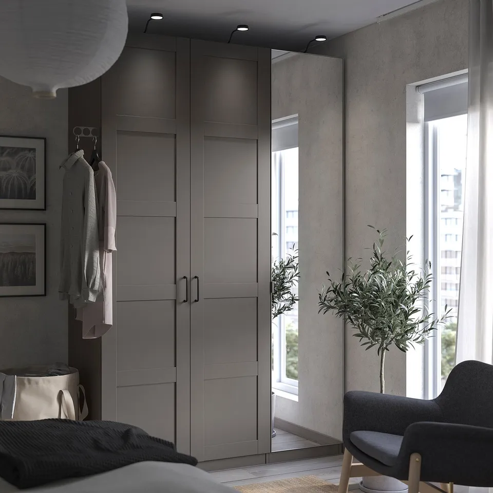 Modern grey PAX wardrobe with mirror doors beside a window, featuring a grey chair and a tall plant, providing a minimalistic bedroom setup.