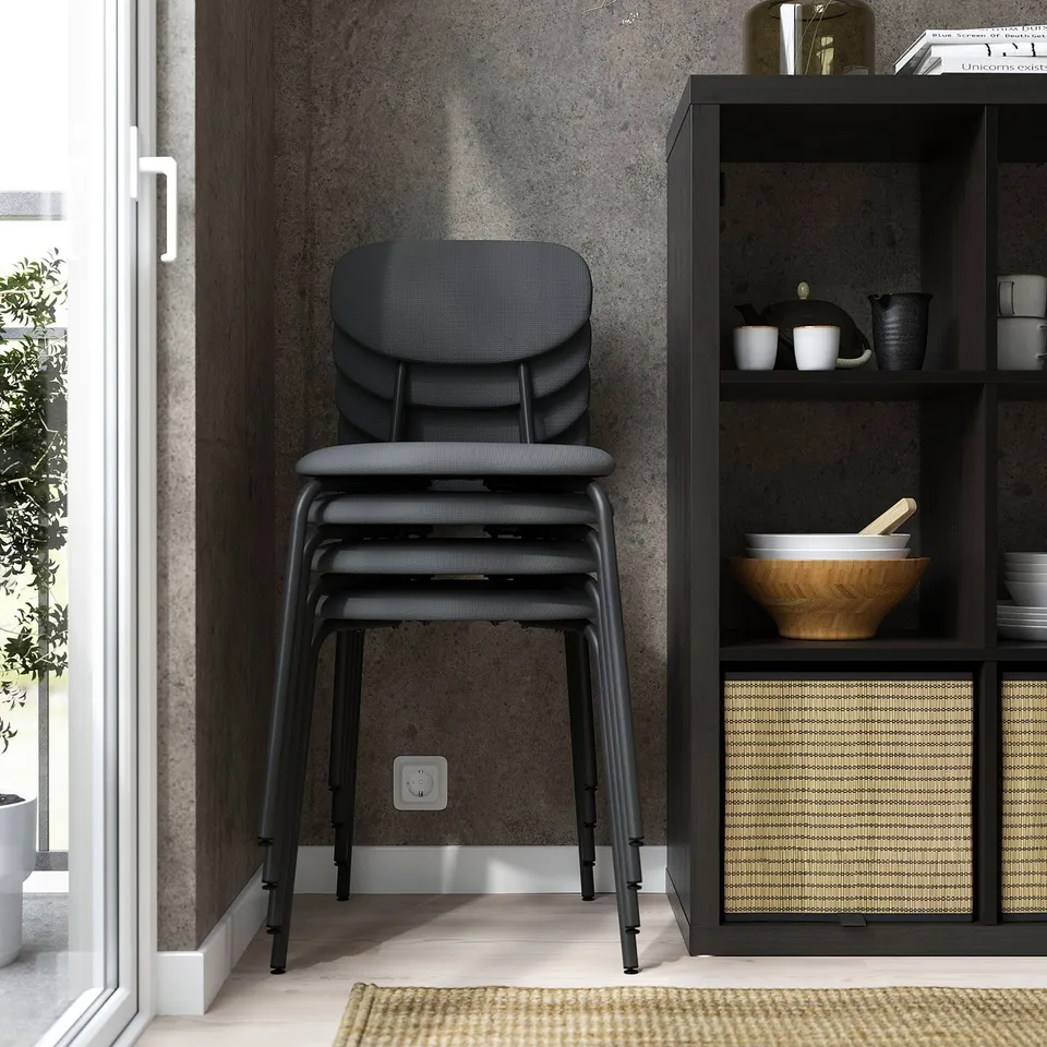 Stacked modern black chairs, arranged neatly against a wall with shelves displaying pottery and a plant, near a door with light streaming in.