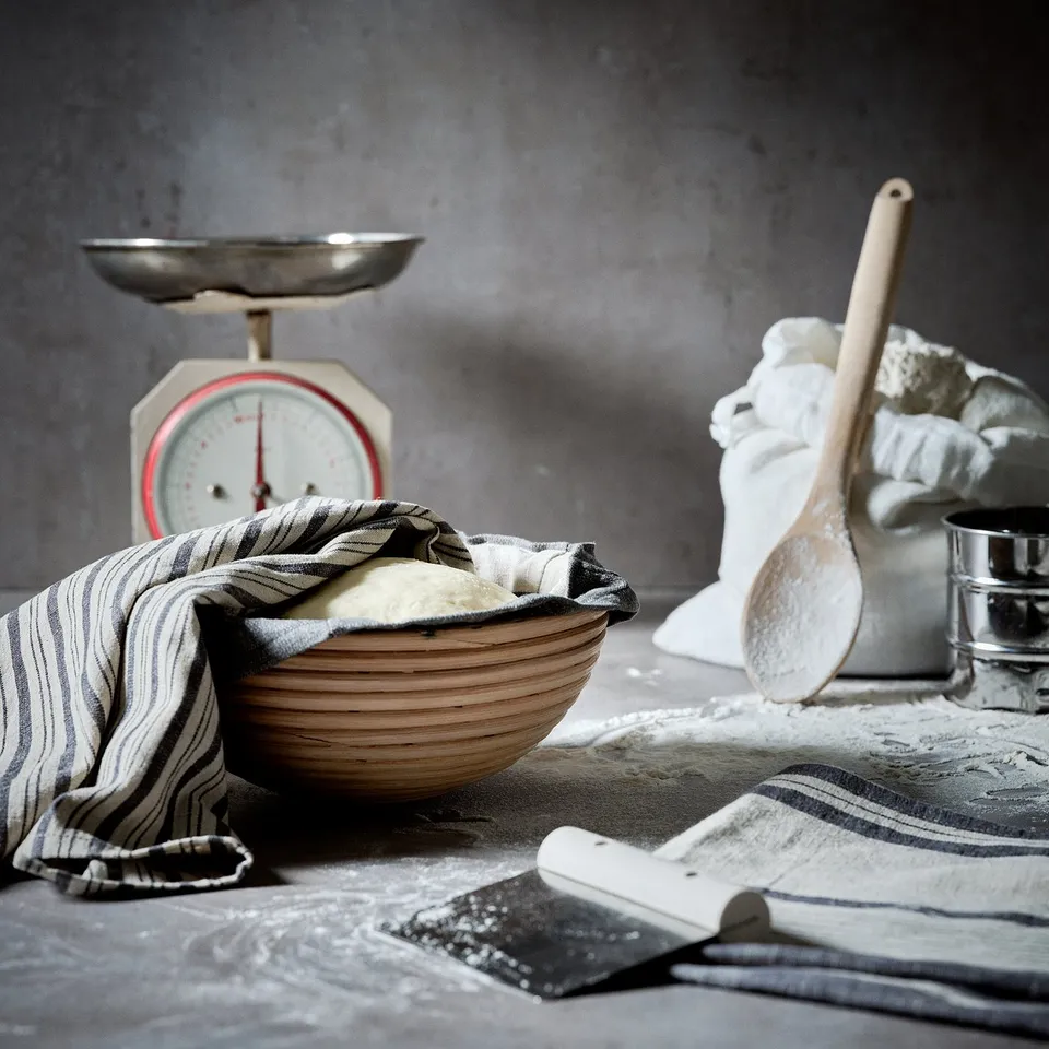 Kitchen scene with dough in basket, scale, spoon, and utensils. Dark grey background with flour.