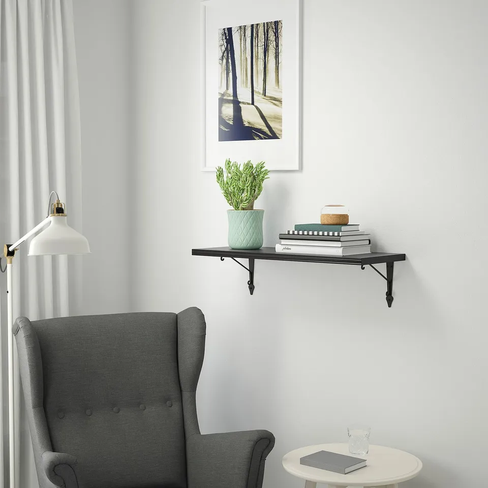Black BERGSHULT shelf with plant and books above grey chair.
