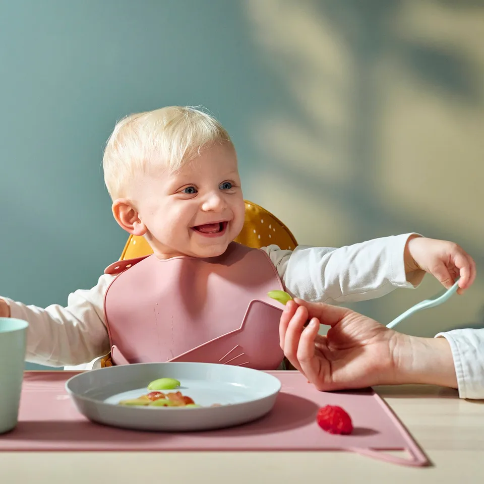 A baby in a high chair wearing a silicone bib with a pocket catches food. An adult feeds the baby with a spoon.