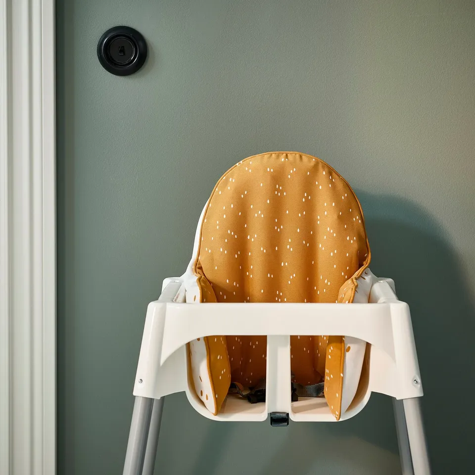 A white high chair with a yellow and white spotted cushion provides stable support for a baby, attached to a green wall.