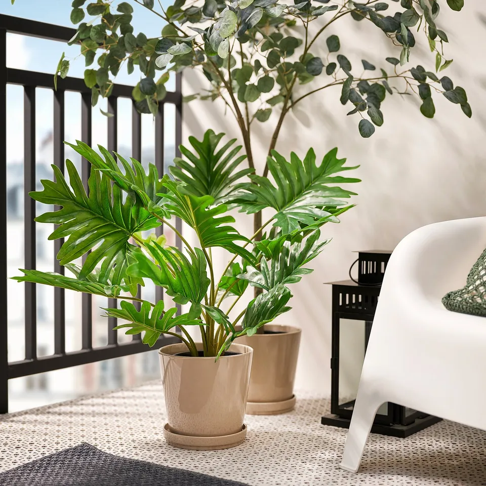Two FEJKA potted plants on a balcony table, bright green leaves in beige pots near a white chair and black lantern.