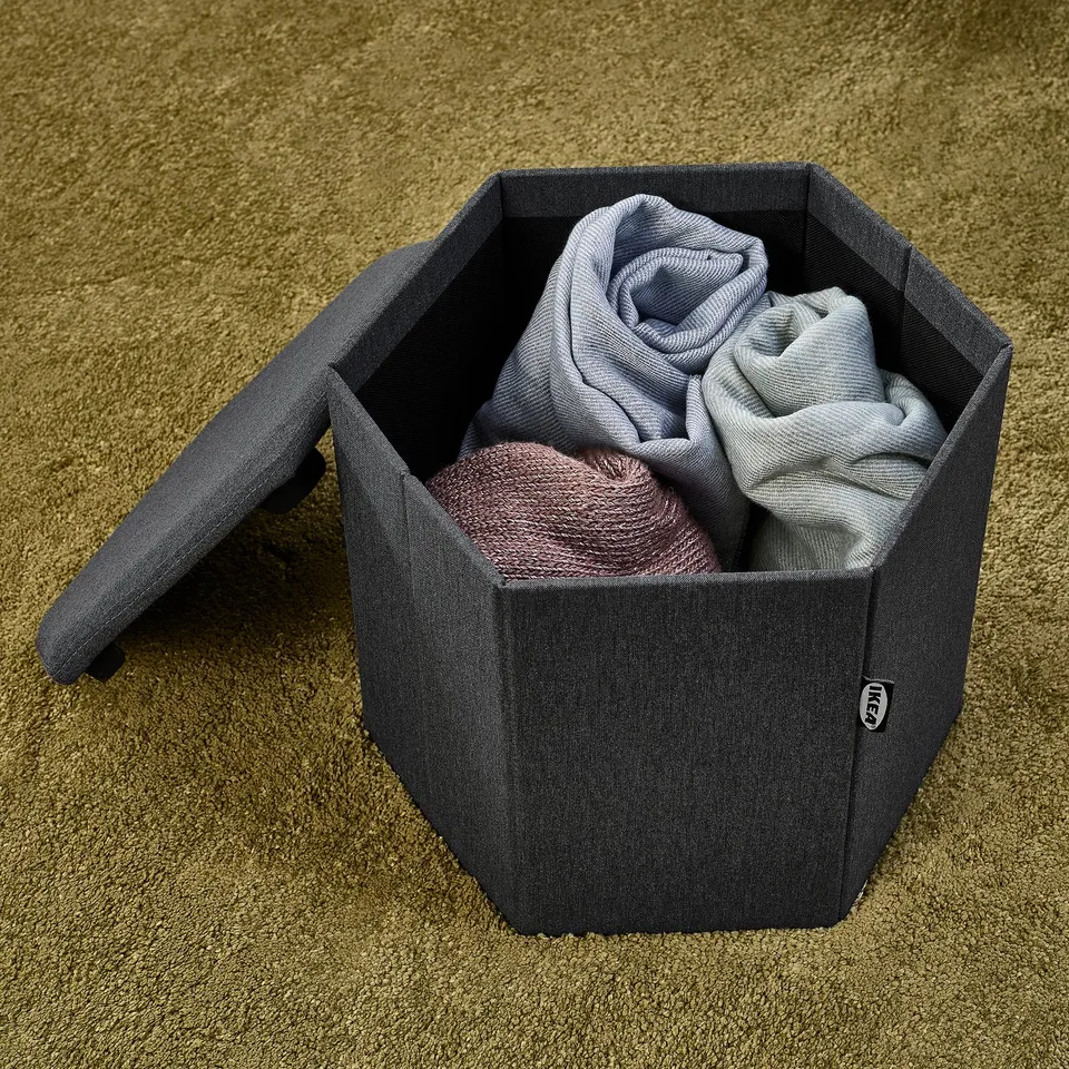 Grey fabric storage box with lid open, revealing folded clothes inside, on green carpet. Extra seat and organiser.