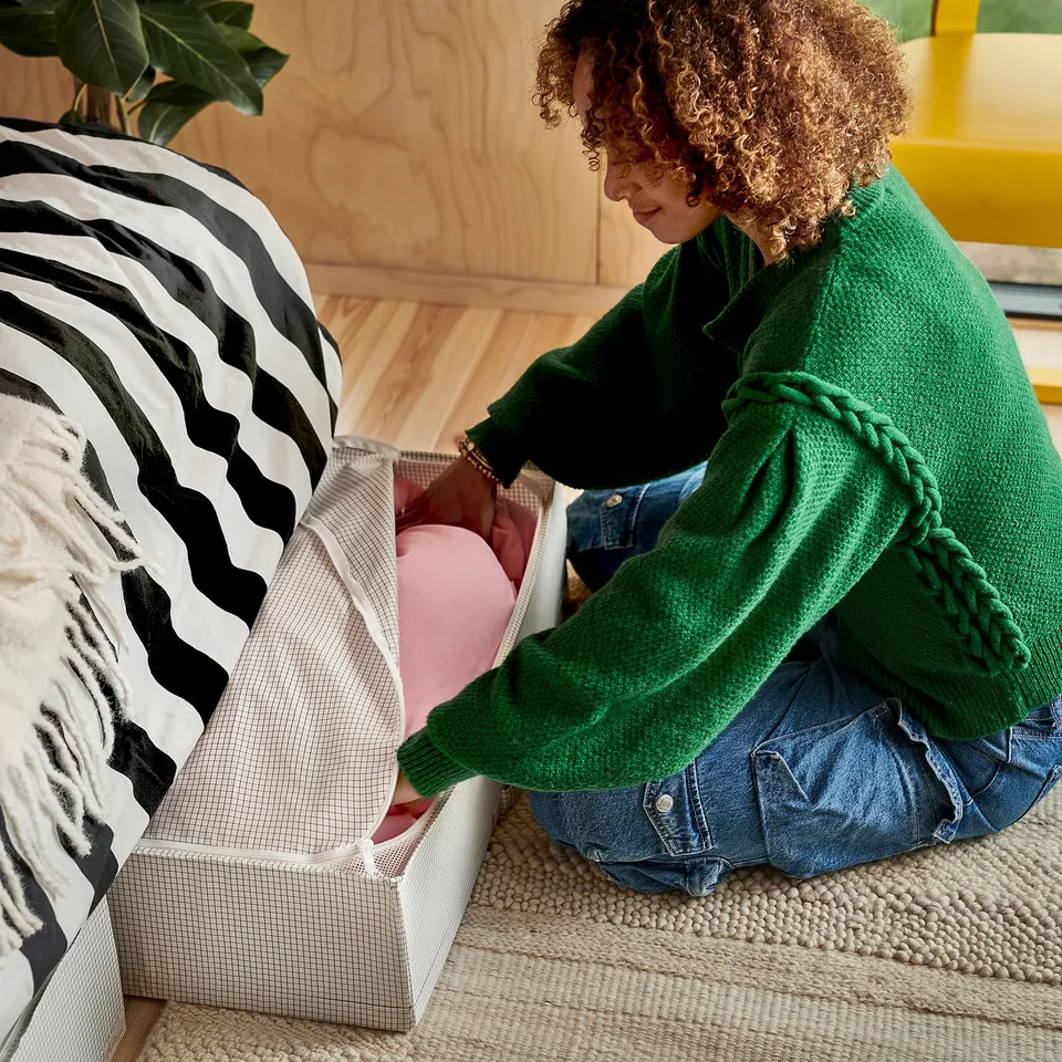 Person arranging cushions in a white basket on a woven rug near a bed with black and white striped pillows.