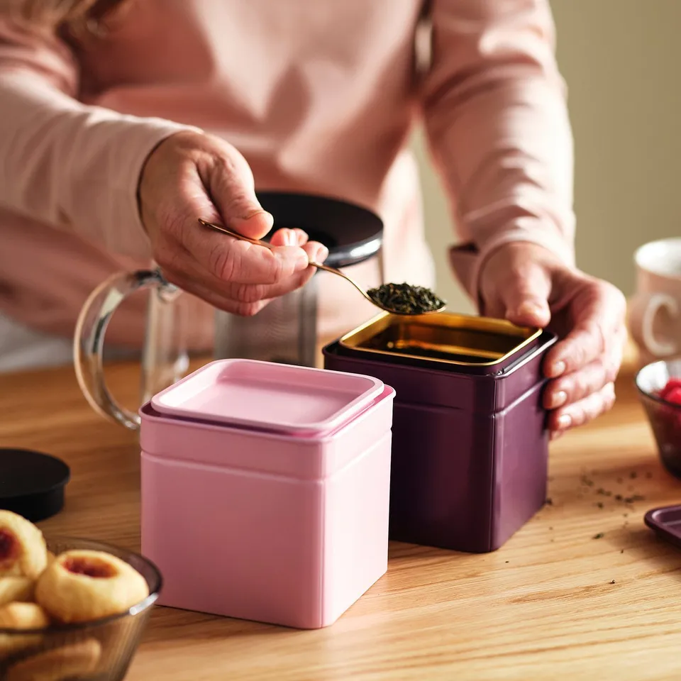 Person scoops tea leaves from purple container. Stackable set for coffee, tea, spices.