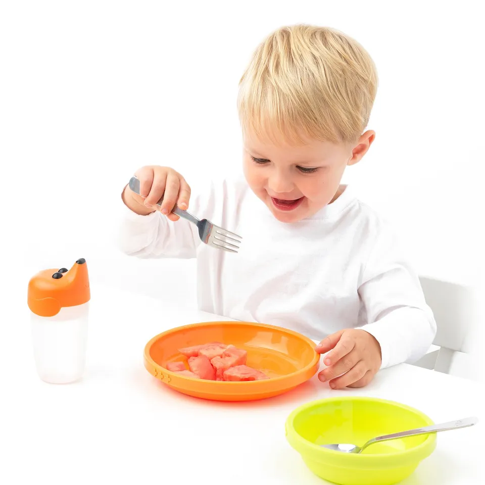 Child eating watermelon with fork, sippy cup, spoon nearby.