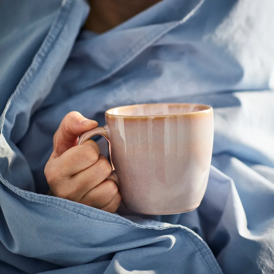 Person holds steaming mug under blue blanket, enjoying a cosy break with FÄRGKLAR mug.