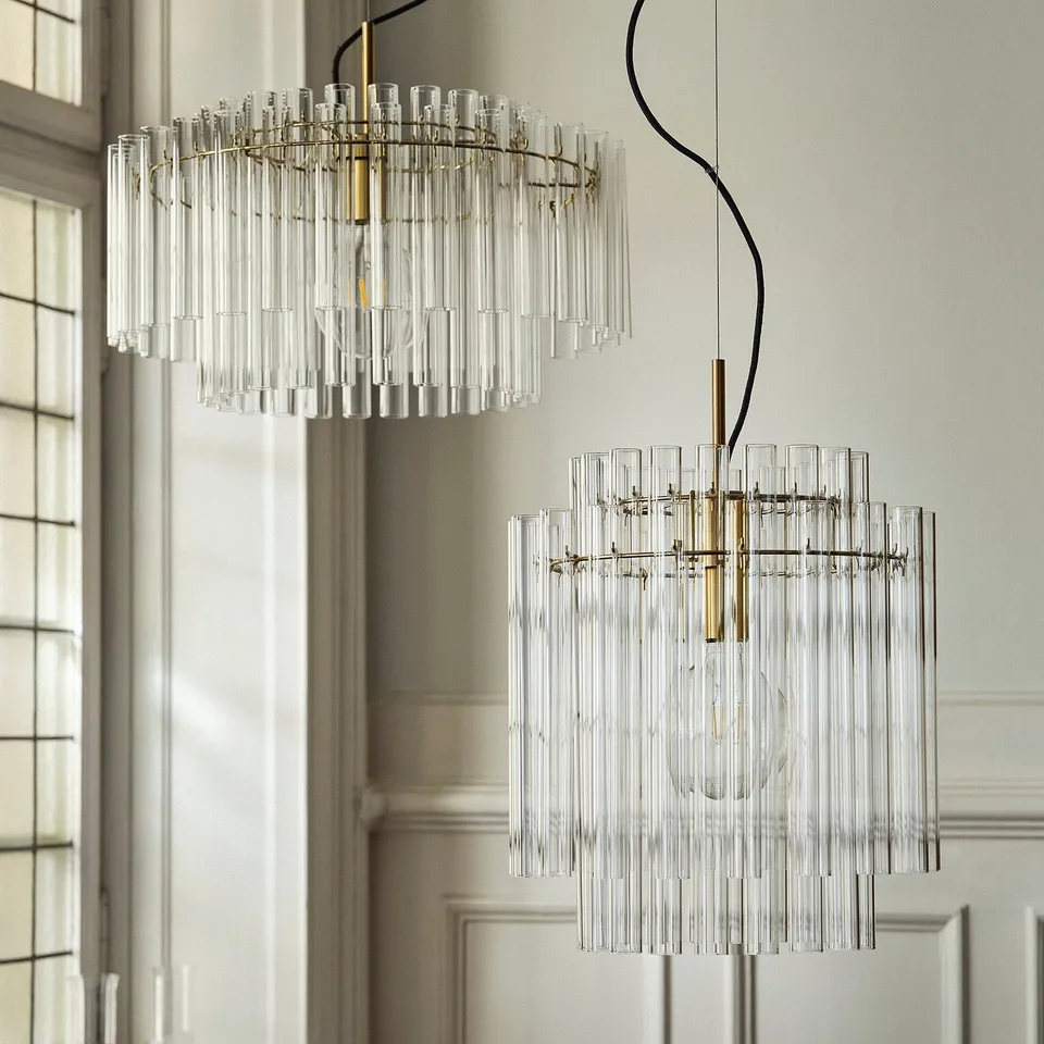 Two STOCKHOLM 2025 chandeliers with clear glass and brass, circular and rectangular, modern and elegant, dominant, brightening room.