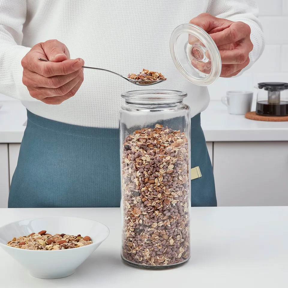 Person spooning cereal from glass jar with lid off onto bowl, showcasing transparent storage.