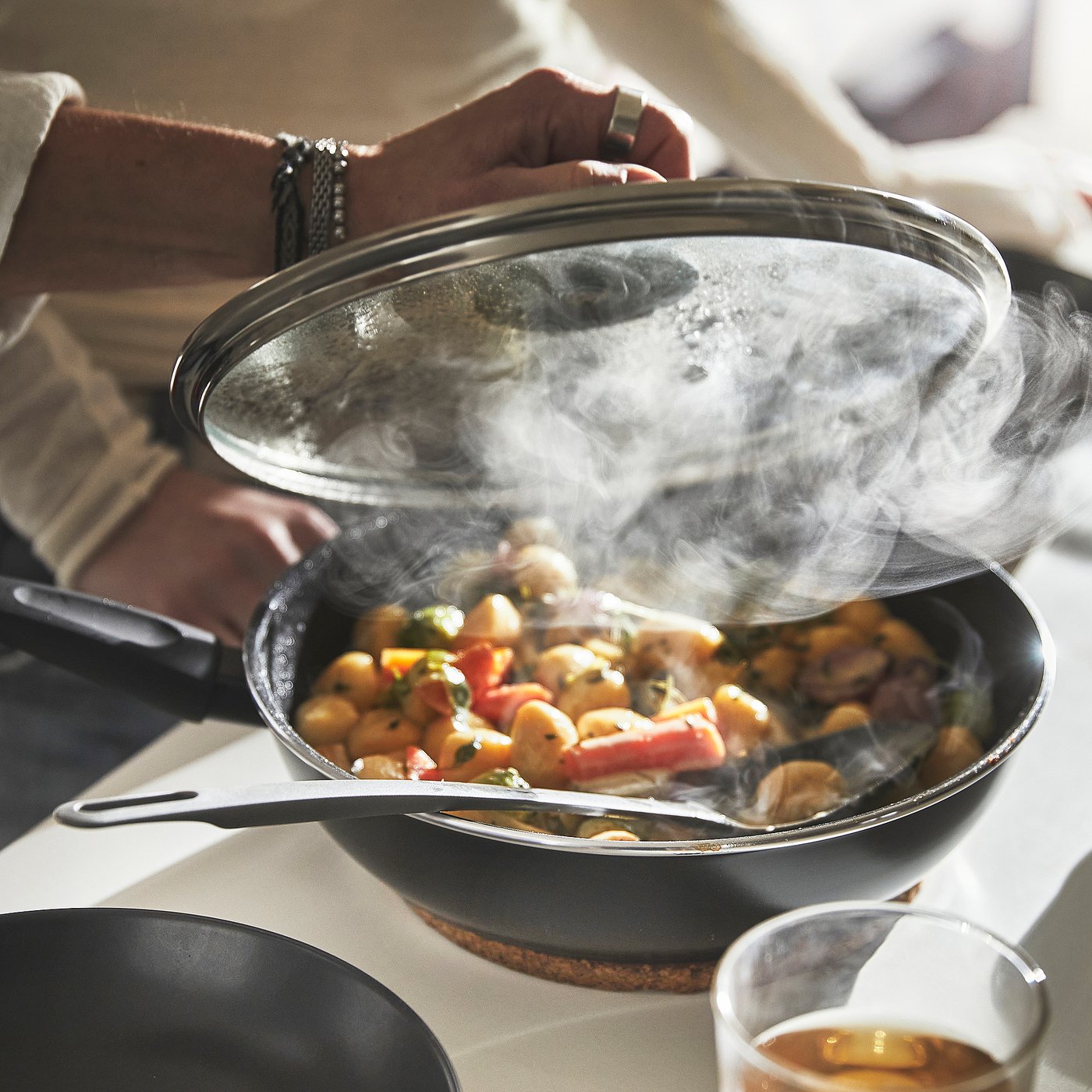 Person lifts lid from sizzling pan, releasing steam. Shows cooking with HEMLAGAD, even heat distribution, steam braising food.
