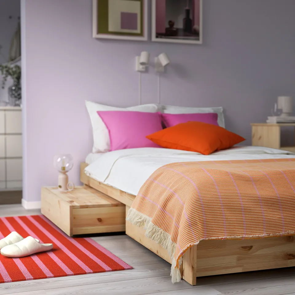 Bed with orange, pink, & white pillows. Striped blanket, wooden nightstands, slippers. Purple walls, art. Cosy, colourful bedroom.