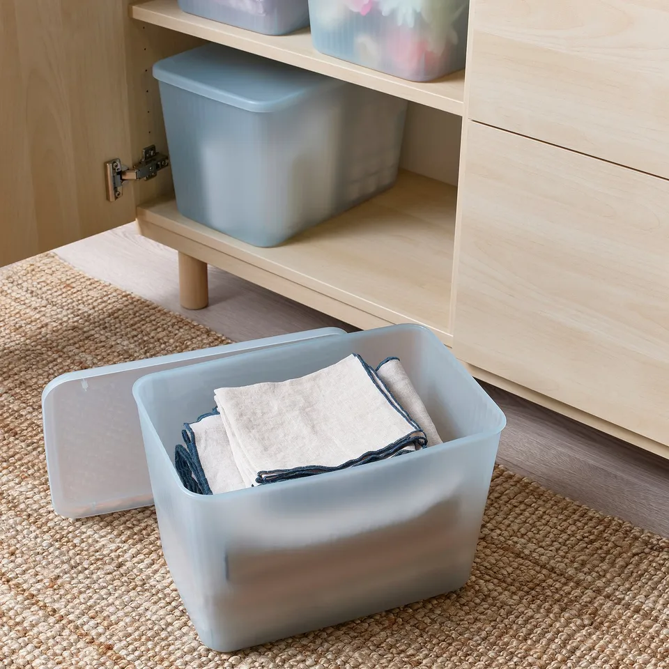 Plastic storage box with lid on rug, organising items next to wooden cabinet.