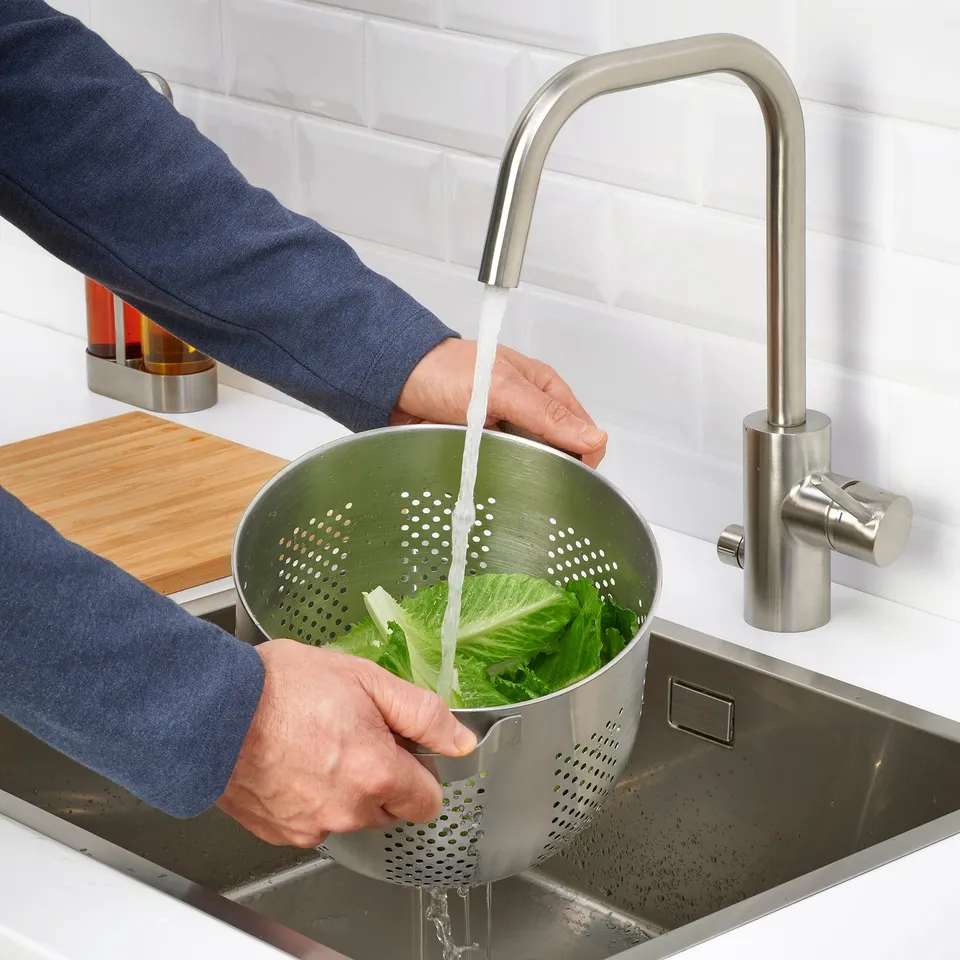 Person washing green leafy vegetables in a metal colander under a kitchen tap.