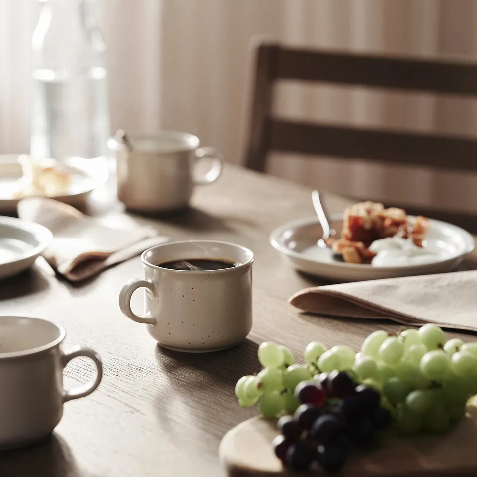 Small cups of DAGGASTER light beige stoneware sit on a wooden table. Theyre surrounded by grapes, plates, and napkins, creating a cosy setting.