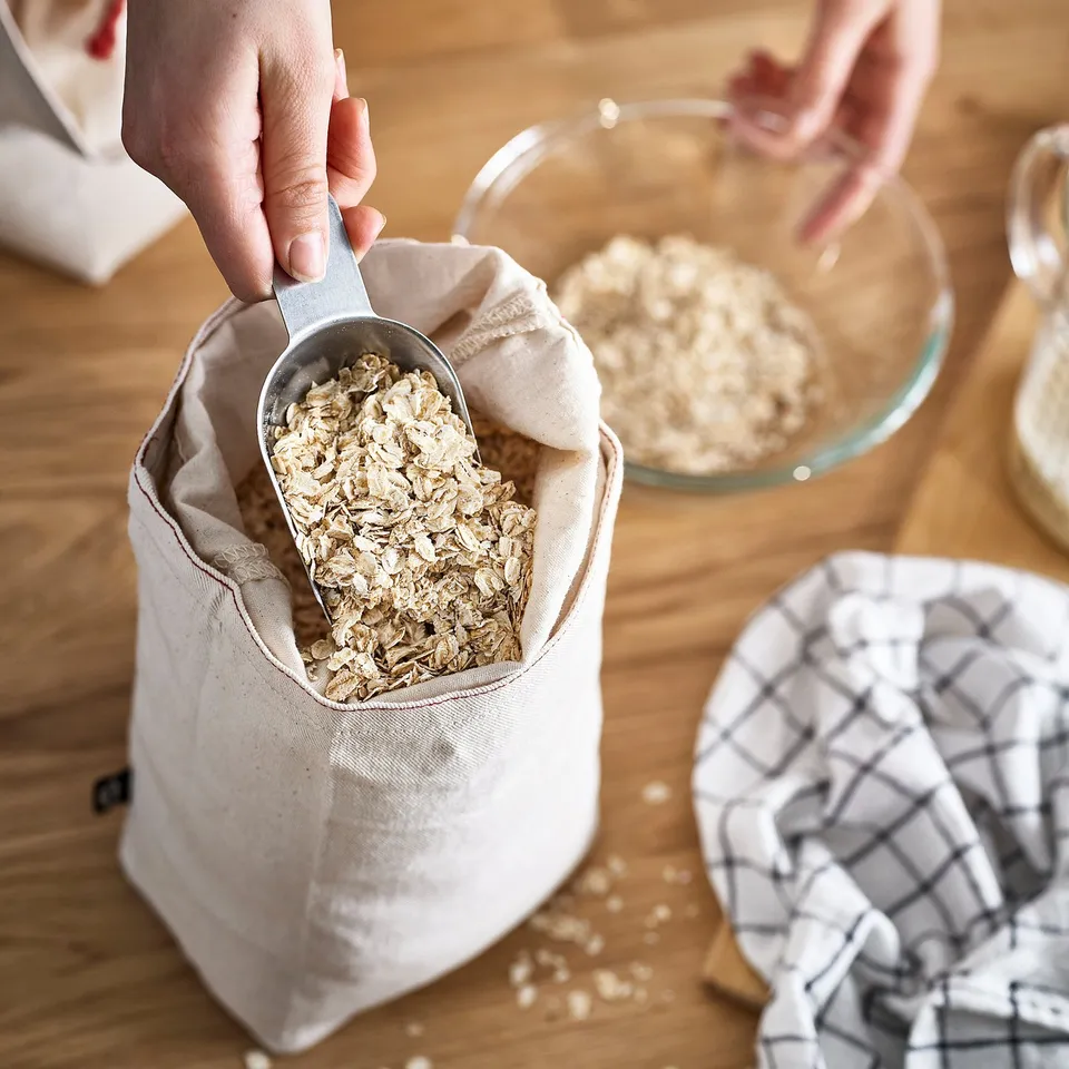 Person scooping oats from a white fabric bag on a wooden table. Other kitchen items nearby.