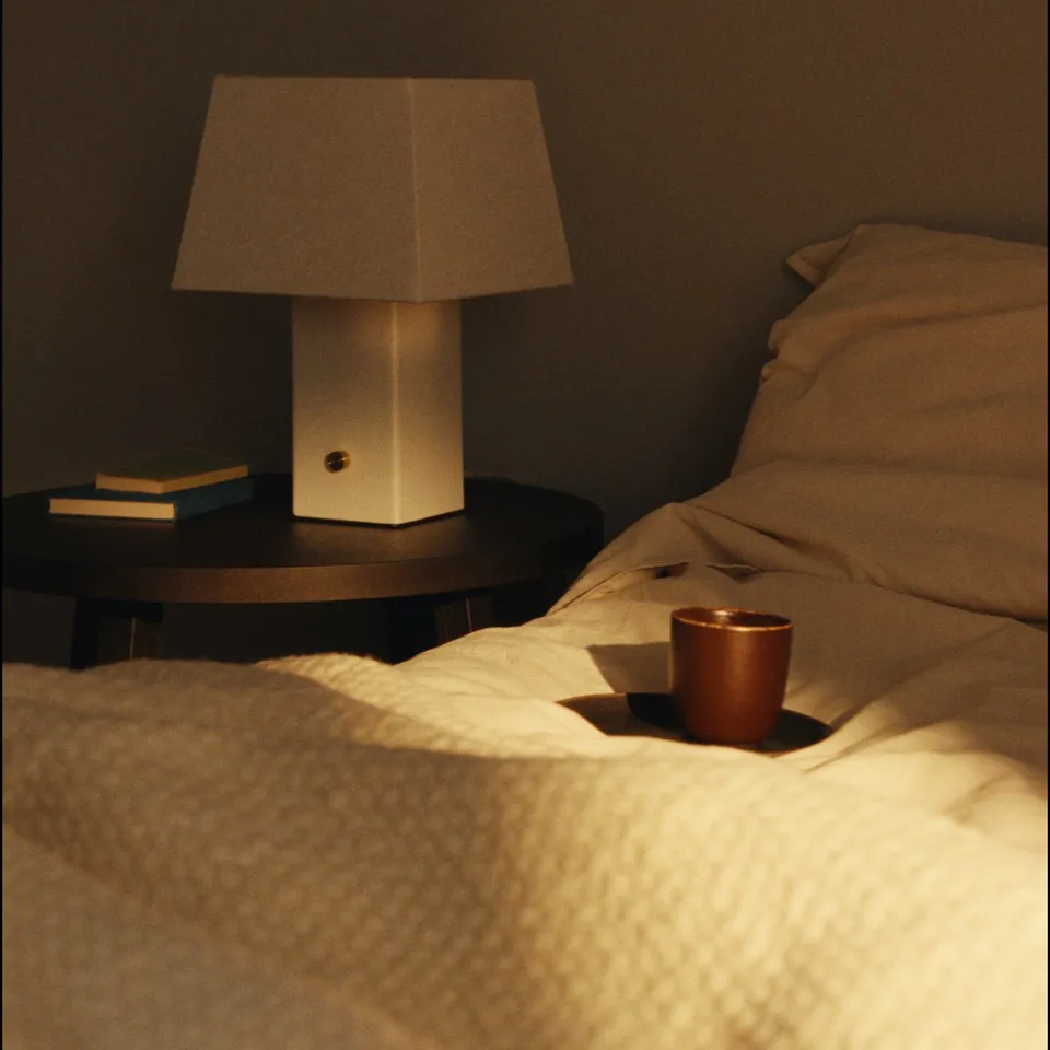 A bedside table with a modern lamp, two stacked books, and a small copper cup on white sheets.