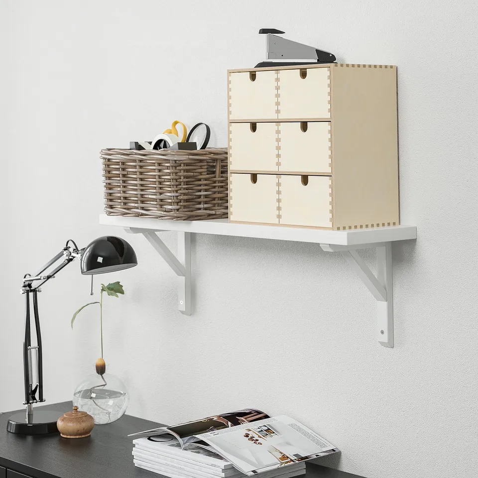 Wall-mounted shelf with boxes and a basket holding office supplies, with a lamp and magazines below.