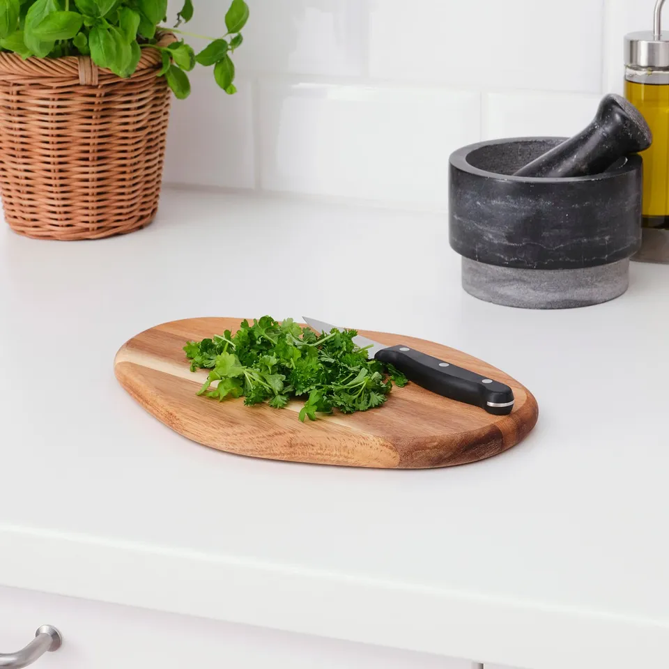 Wooden chopping board with knife and parsley on white countertop, beside a basket with plant and mortar.