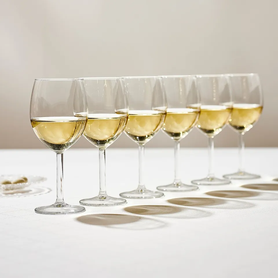 Six wine glasses filled with white wine on white tablecloth.