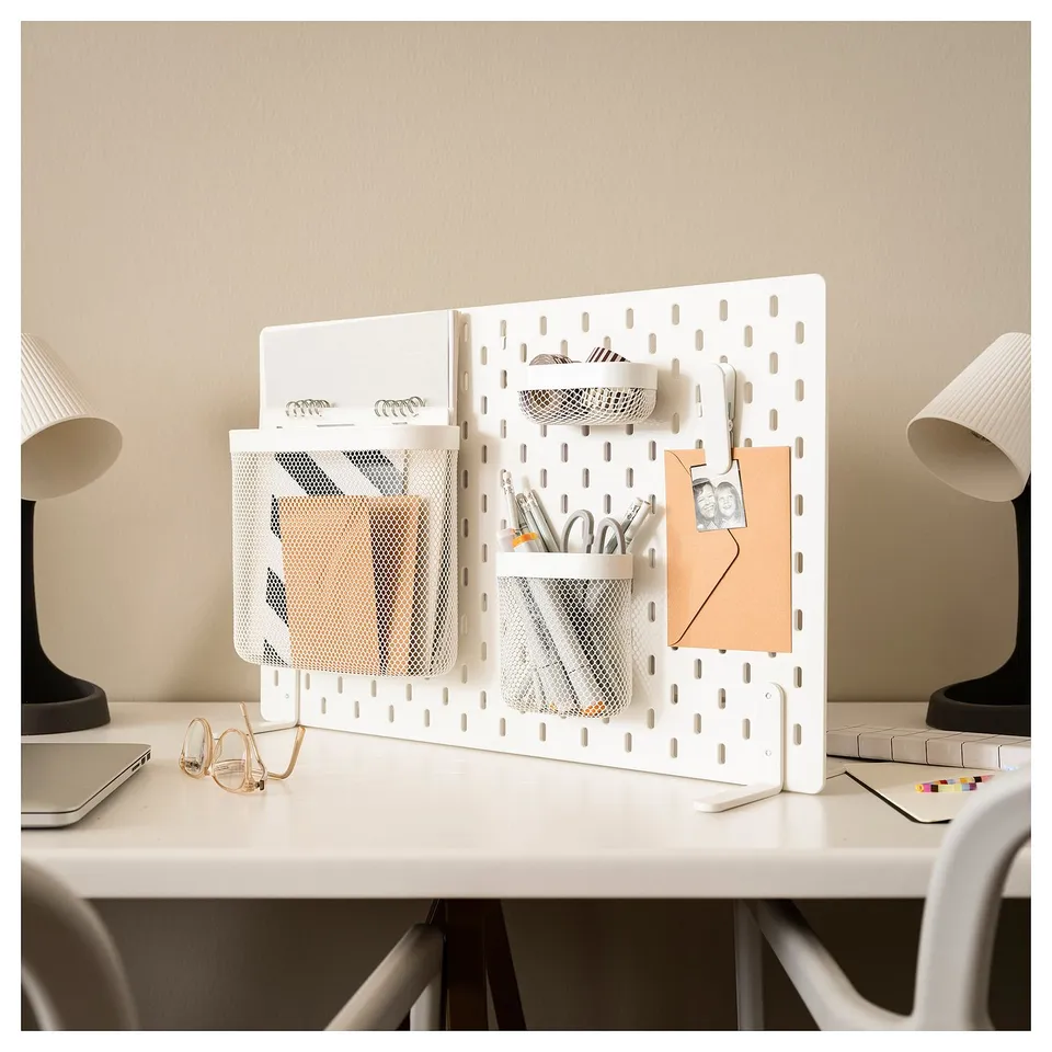 A white SKÅDIS pegboard organiser with mesh storage pockets on a white desk. It holds office supplies and a laptop, flanked by white lamps.