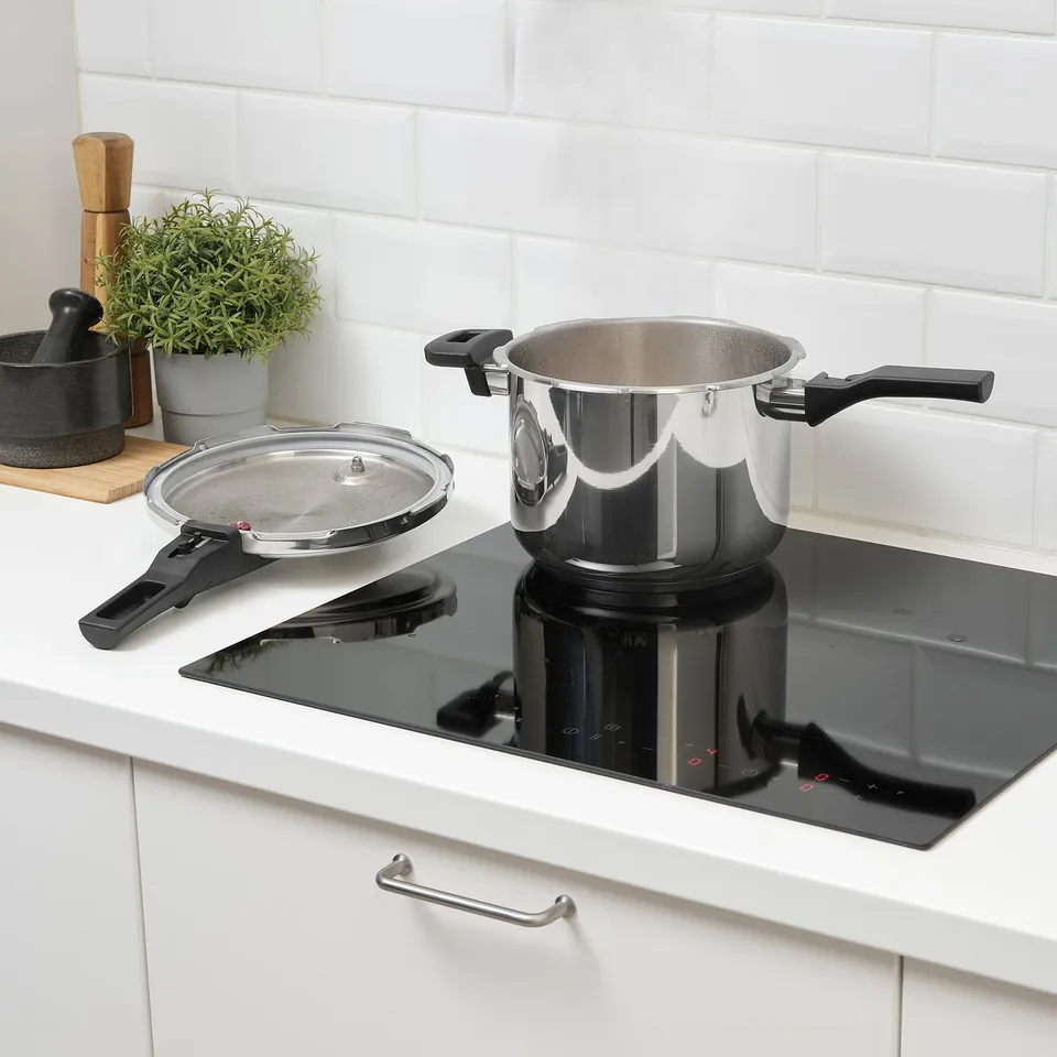 Stainless steel pressure cooker on black induction hob with lid off, showcasing handles and safety regulator.