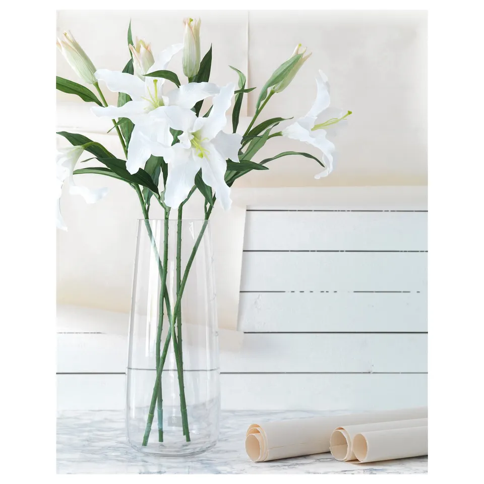 A clear vase with white artificial lilies, green stems, and leaves. Set on a white marble surface, beside rolled papers.