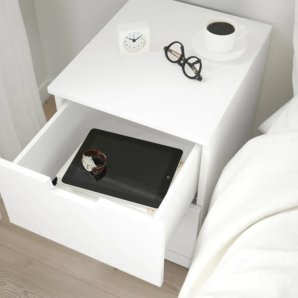 White bedside table with open drawer showing a tablet and watch, glasses, and coffee cup on top.