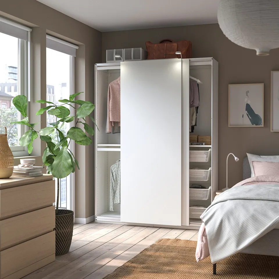 Modern bedroom with open wardrobe showing clothes, shelves, and storage baskets. PAX wardrobe in white, wood floors, and plant near window.