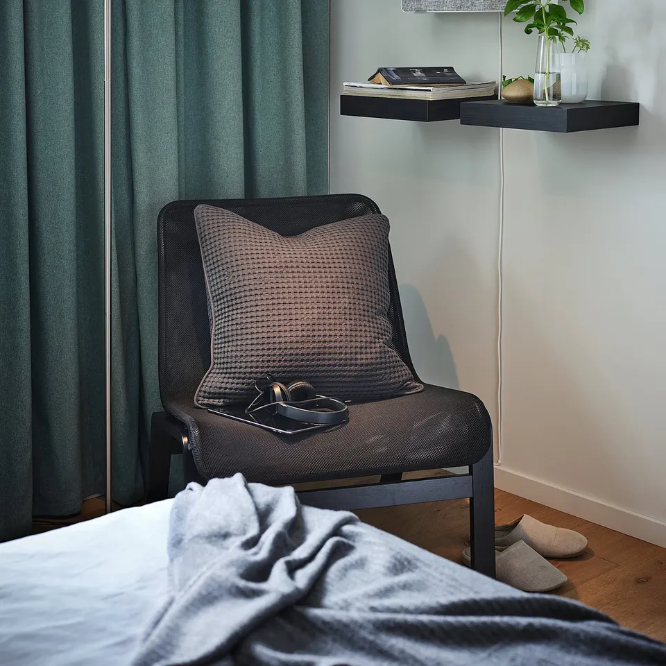 Modern bedroom with grey armchair, dark curtains, floating shelves with plants, books, and lamp.