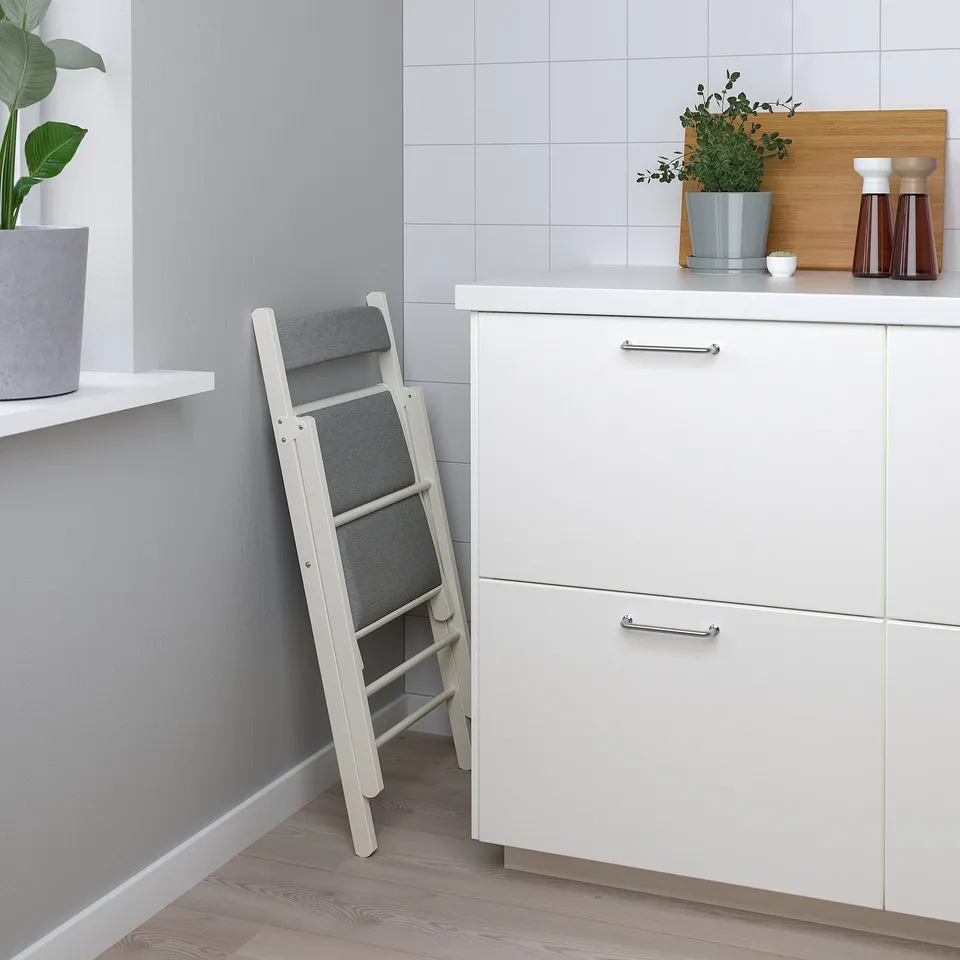 Small white kitchen with foldable step stool, grey cushions, plants, and wooden countertop.