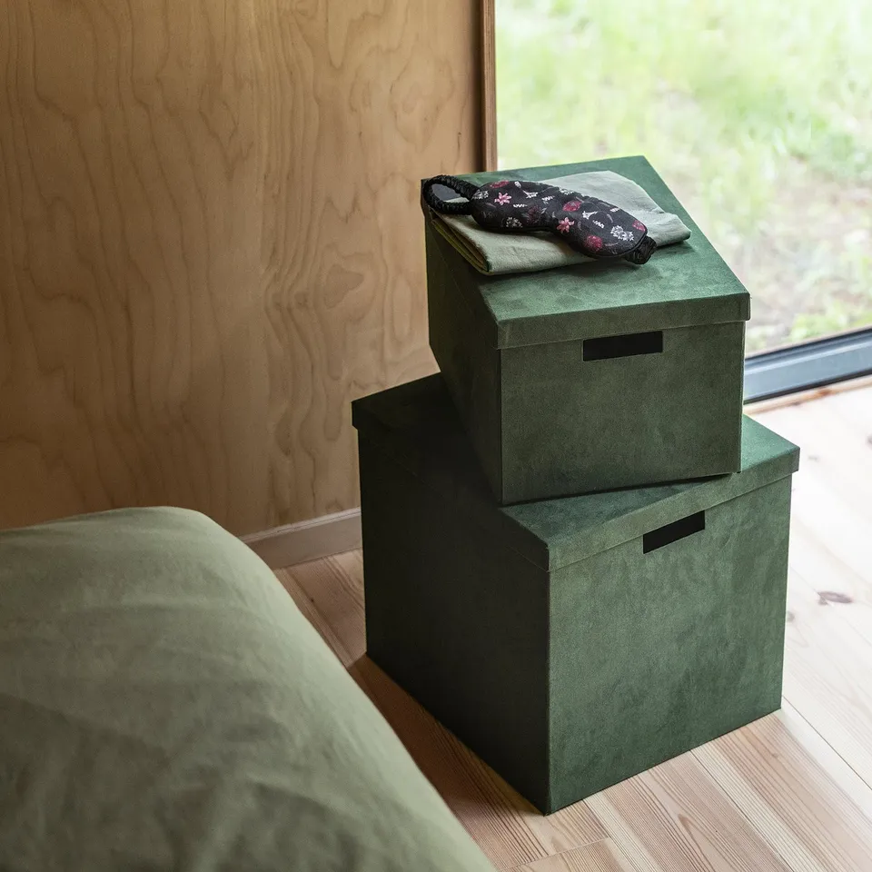Green velvet GJÄTTA boxes with clothes. Stacked near window, top has floral item.