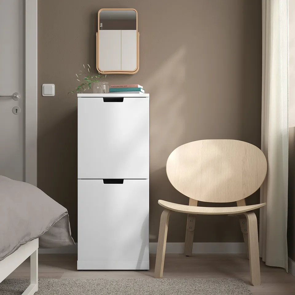 A modern white NORDLI chest between bed & window, topped with decor, beside a chair. Ideal for small rooms.