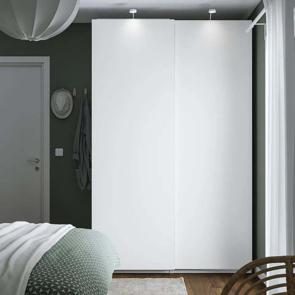 Minimalist bedroom with large white PAX wardrobe, grey door, hanging hat, and bed with green blanket.