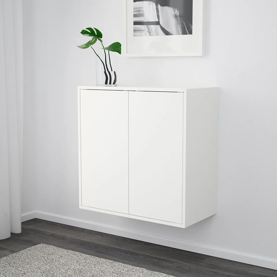 White wall-mounted cabinet with minimalist design, featuring two doors and a modern storage solution. A plant on top adds a touch of decor.
