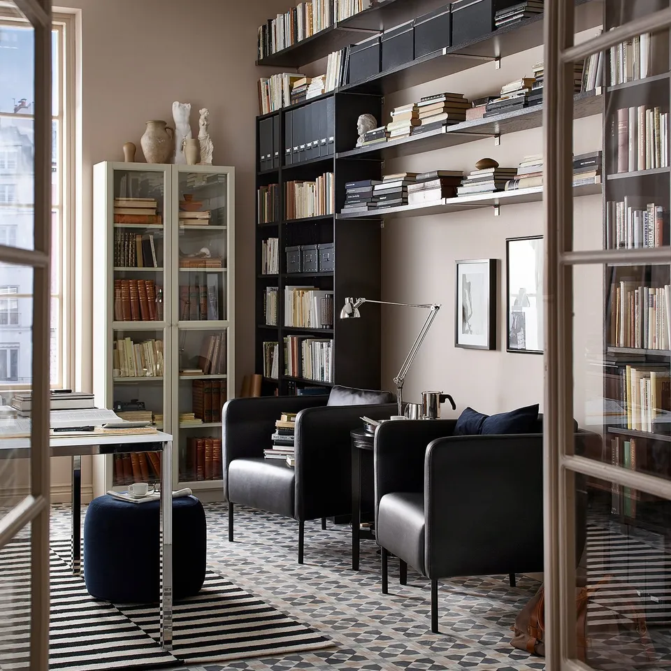 A stylish lounge with black EKERÖ armchairs, bookshelves, and a geometric rug. Large windows let in natural light.
