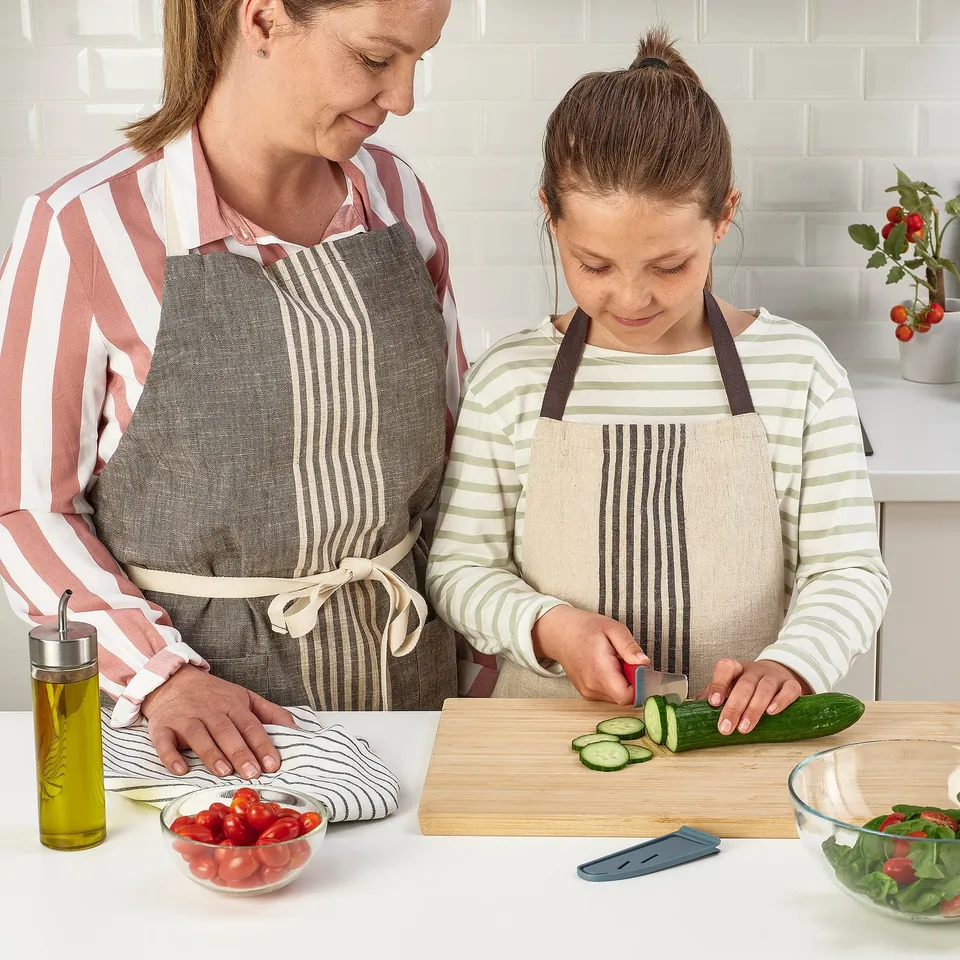 Mother and daughter cooking together, using child-safe knives to slice cucumber and tomatoes on a kitchen counter.