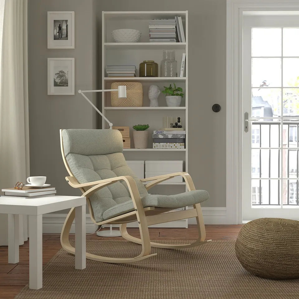 Modern living room with grey rocker chair, matching ottoman, white side table, bookshelf with decor, and large window.